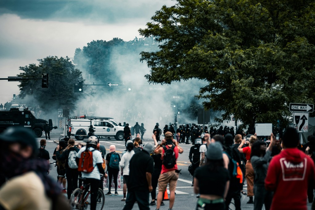 A crowd standing back from a large group of emergency or law enforcement personnel further down a road while smoke rises into the sky.