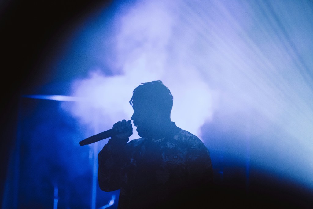 A silhouette of a young person framed in light and haze with a purple shade to the whole image. The person holds a microphone and appears to be in the middle of a song.